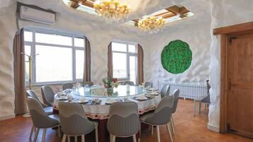 A round dining table with chairs, a chandelier, and a green wall decoration in a room with wooden floors and large windows.