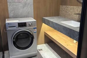 A compact laundry area with a washing machine, a sink, and a wooden shelf. - Forest Snow Hotel (Genhe)