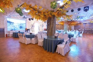 Un salón de banquetes con mesas y sillas redondas, un centro de mesa con un gran árbol y un escenario con un telón de fondo.