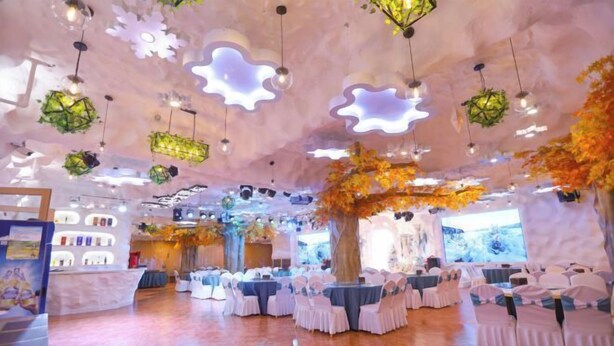 A spacious hall with round tables and chairs, a large tree centerpiece, and hanging light fixtures. - Forest Snow Hotel (Genhe)