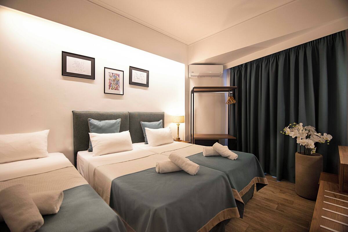 Une chambre d’hôtel avec deux lits, une grande fenêtre avec des rideaux, un appareil de climatisation et un vase avec des fleurs.