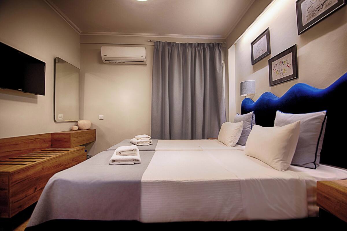 Une chambre d’hôtel avec un lit, un banc en bois, une télévision, un climatiseur et des œuvres d’art accrochées au mur.