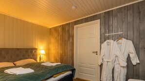 Un dormitorio con una cama, una mesita de noche con lámpara, y una puerta con dos batas blancas colgadas en un perchero.