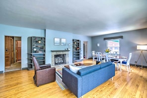 A spacious living room with a fireplace, a blue sofa, wooden flooring, and a dining area with white chairs. - Sugar Shores ~ 3 Mi to Indiana Dunes Nat'l Park (Chesterton)