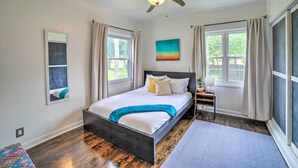 Une chambre à coucher avec un lit, un ventilateur de plafond, une fenêtre avec des rideaux, une table de chevet et un tableau au mur.