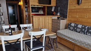 A cozy cabin interior with a dining table set for four, a sofa with patterned cushions, a wooden bar area, and a television mounted above the bar. - Comfortable Apartment, Ski-in/Ski-out, Balcony, Wifi (Les Arcs 1800)