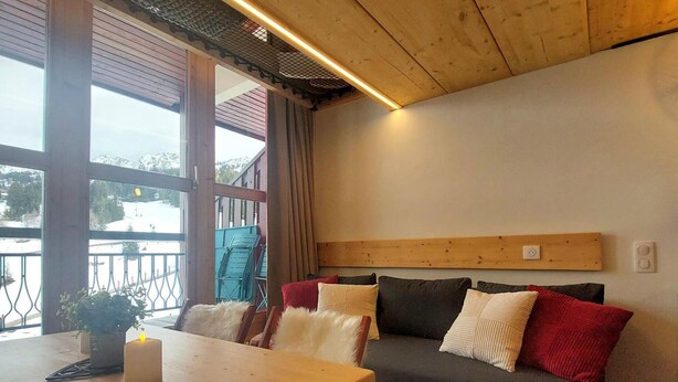 A cozy room with a wooden table, a sofa with cushions, and a view of the outdoors through large windows. - Mezzanine Duplex 5 pers. · Ski-in/out · Mont Blanc View (Les Arcs 1800)