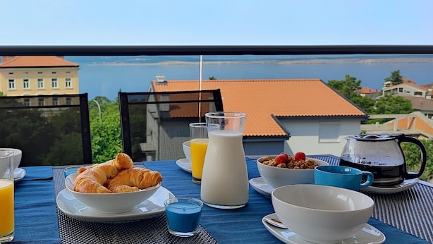A breakfast table with croissants, fruit, juice, and milk, overlooking a scenic view of buildings and water.