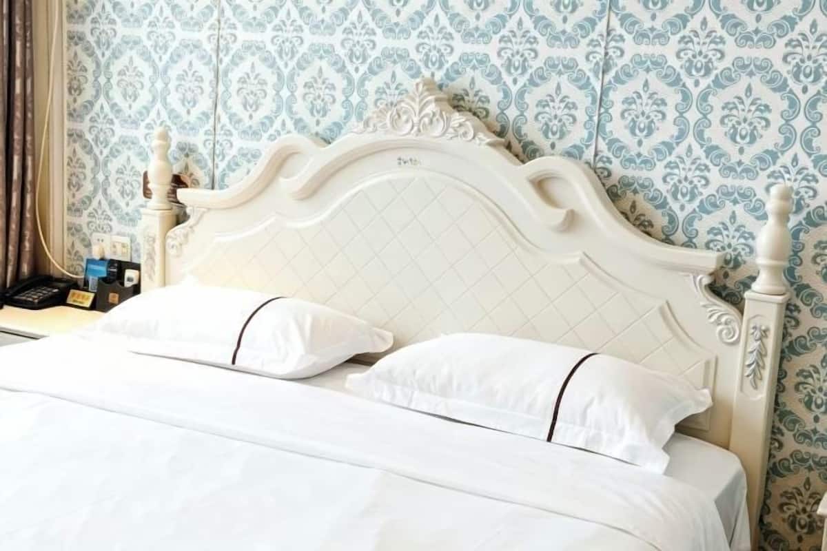 A bed with a white headboard and two white pillows, a bedside table with a phone and a bottle, and patterned wallpaper.