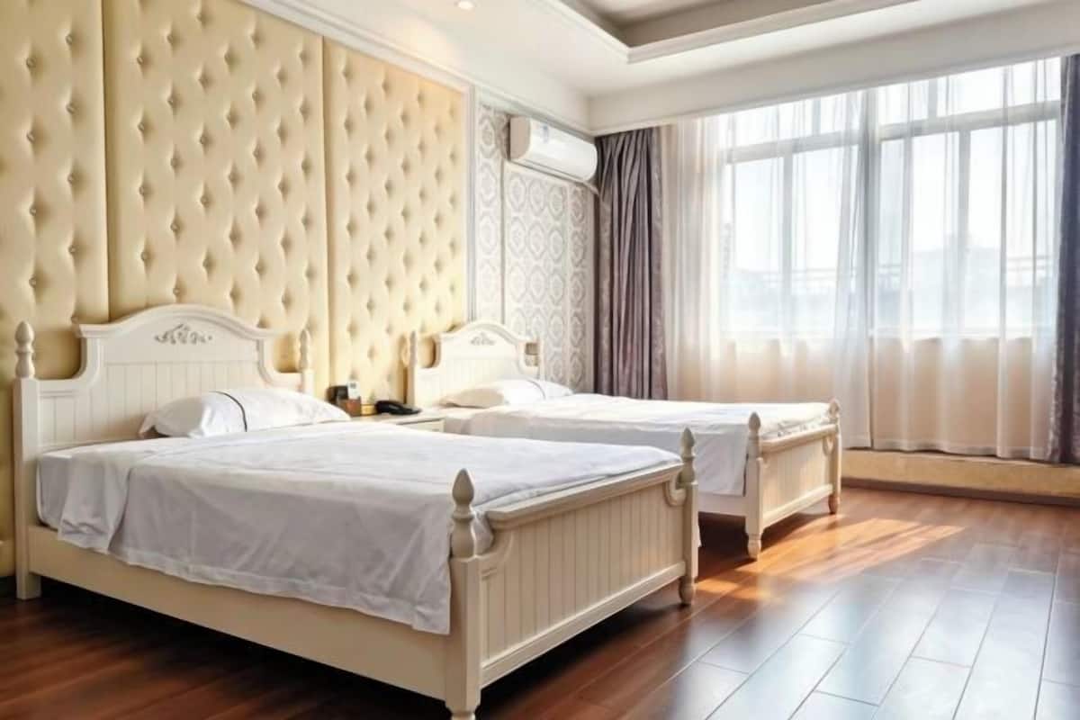 A hotel room with two beds, a tufted headboard, a large window with curtains, and a wooden floor.