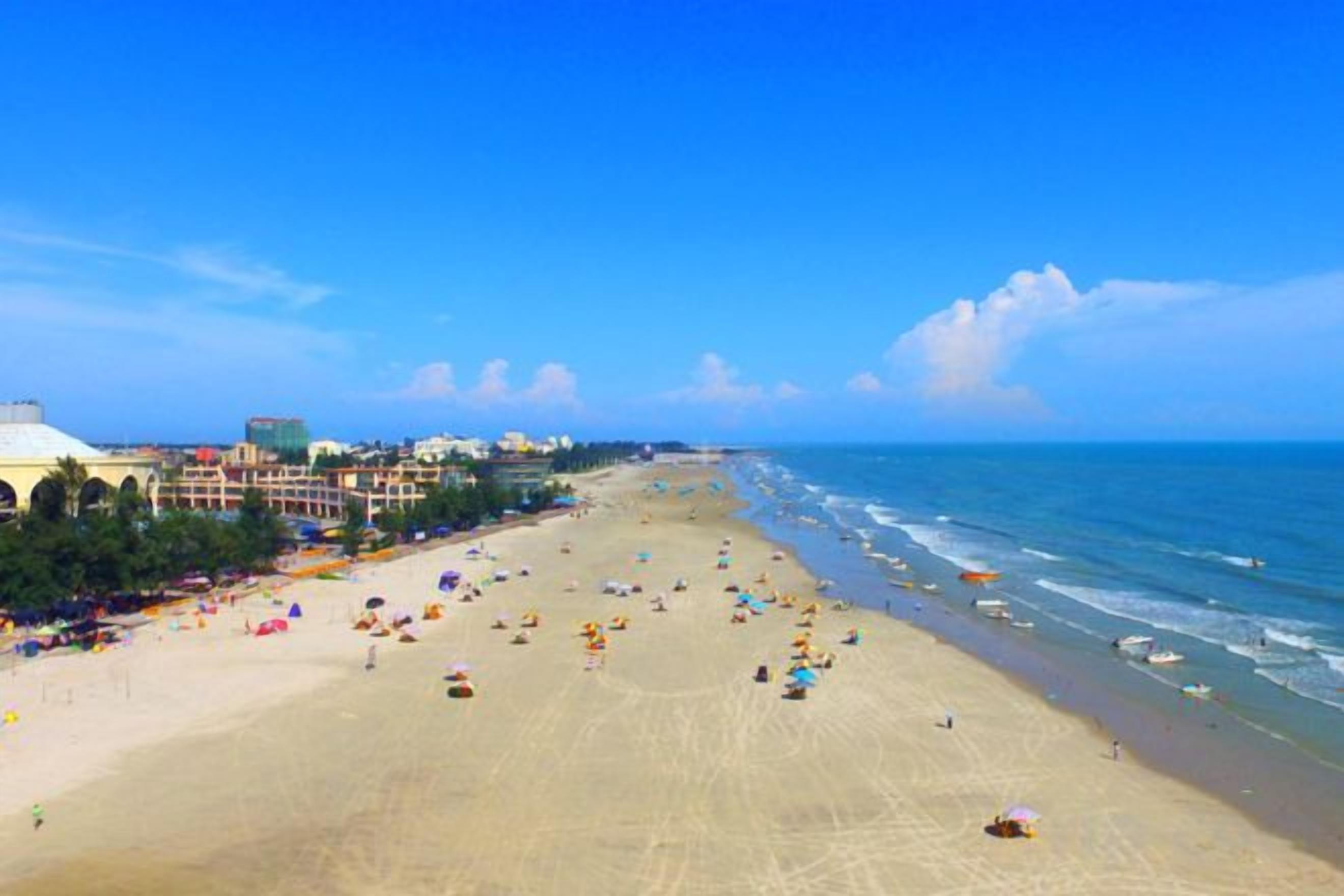 A beach with a long stretch of sand, a few umbrellas, and buildings in the background.