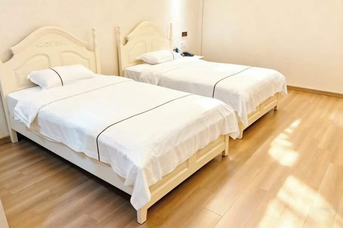 Two white beds with headboards, wooden flooring, and a light switch on the wall.