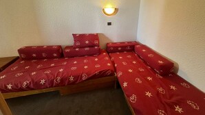 Una habitación pequeña y acogedora con cama y ropa de cama roja estampada con motivos florales, una lámpara de pared y una pared lisa.