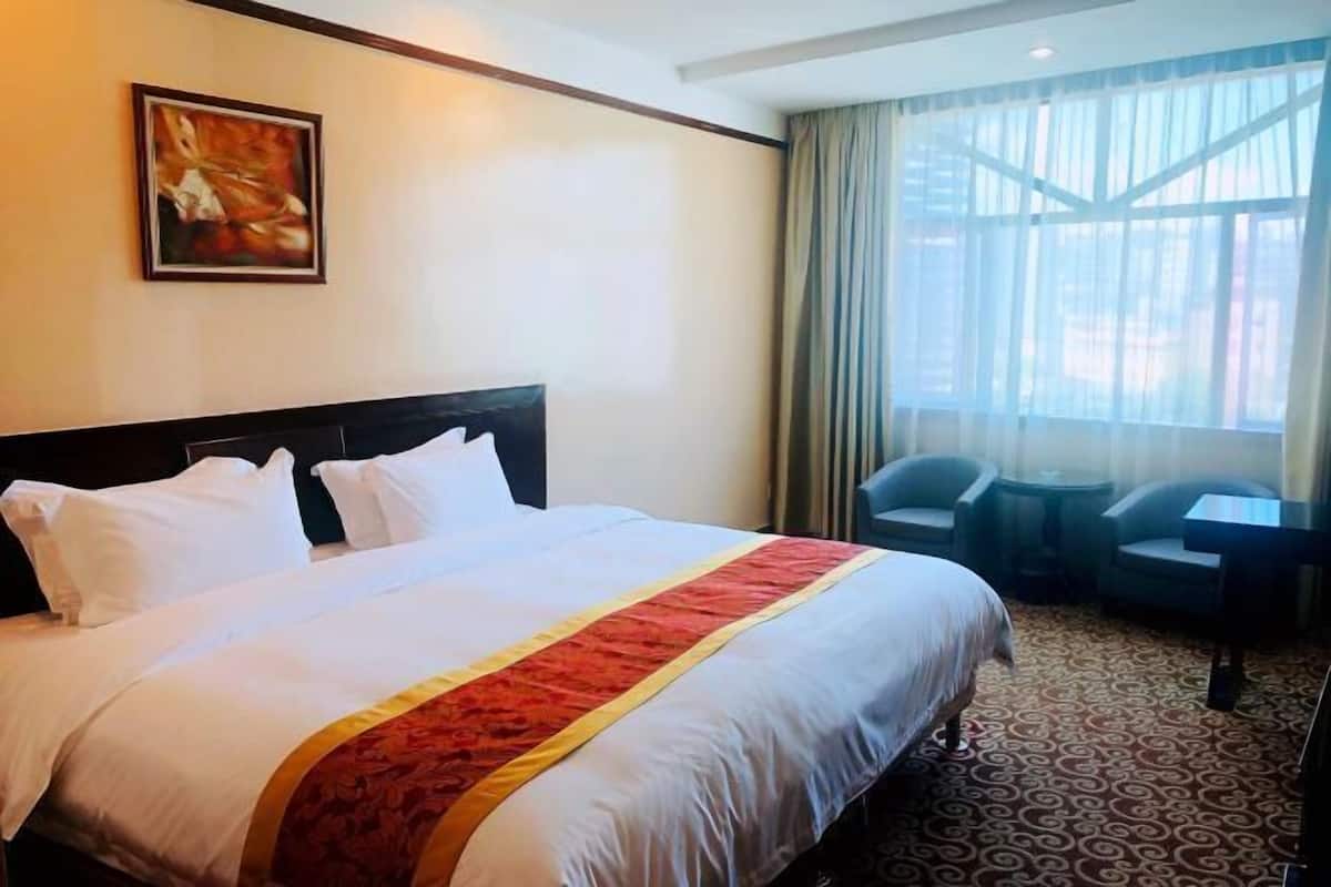 A hotel room with a large bed, a painting on the wall, a window with curtains, and a small table with chairs.