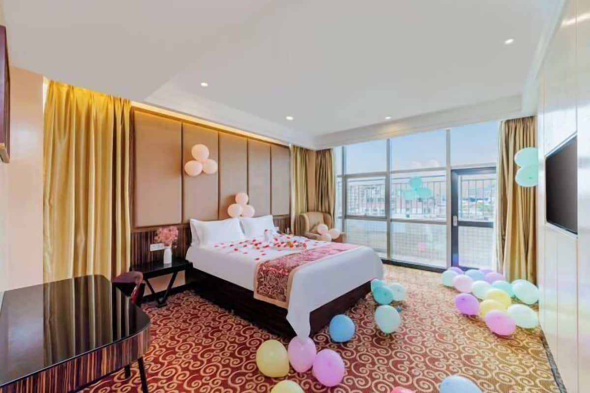 A hotel room with a large bed, a desk, a chair, a TV, and a view of the city through large windows.