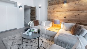 Ett modernt vardagsrum med en grå soffa, ett soffbord med en växt och en flaska, en väggpanel av trä samt ett tak med synliga takbjälkar och lampor.