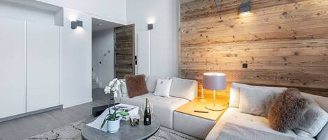 Una sala de estar moderna con un sofá gris, una mesa de centro con una planta y una botella, una pared de madera, y un techo con vigas y luces a la vista.