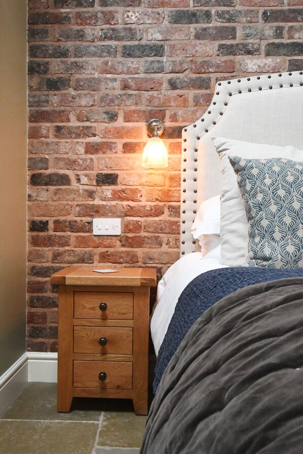 Une chambre avec un mur en briques, une table de chevet en bois dotée de tiroirs, un lit avec une tête de lit et un luminaire fixé au mur.