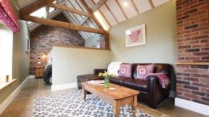 A cozy living room with a brown leather sofa, a wooden coffee table, and a patterned rug. A brick wall and a wooden ceiling beam are visible in the background. - Boutique Farm stay cottage in Worcestershire - Hot Tub / Dog Friendly (worcester)