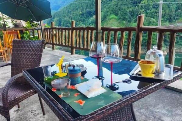 Una mesa dispuesta con copas de vino, un cuenco amarillo y un mantel individual verde en un balcón con baranda de madera y vista a un paisaje verde.