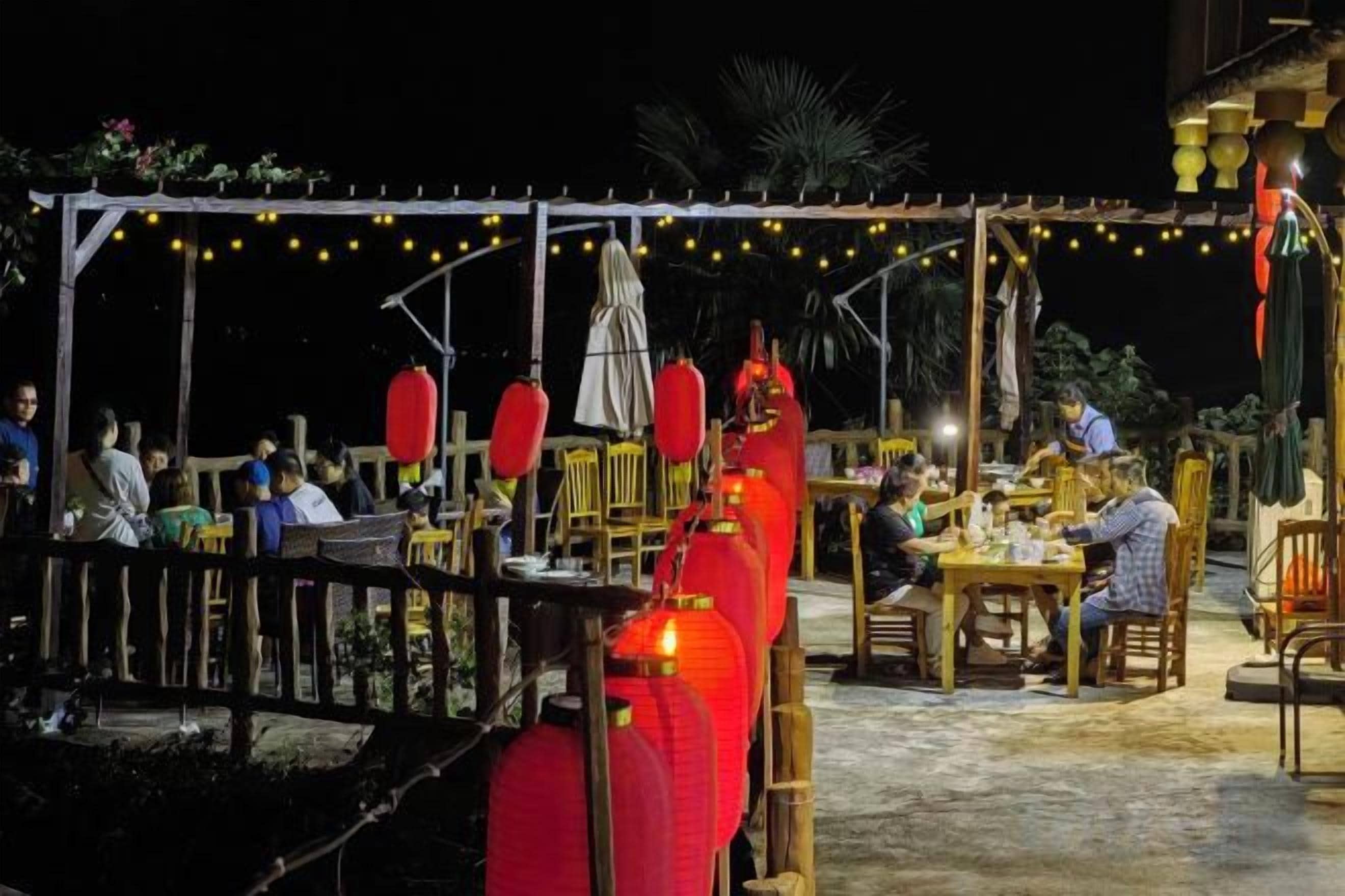 An outdoor dining area with tables and chairs, string lights, and red lanterns.