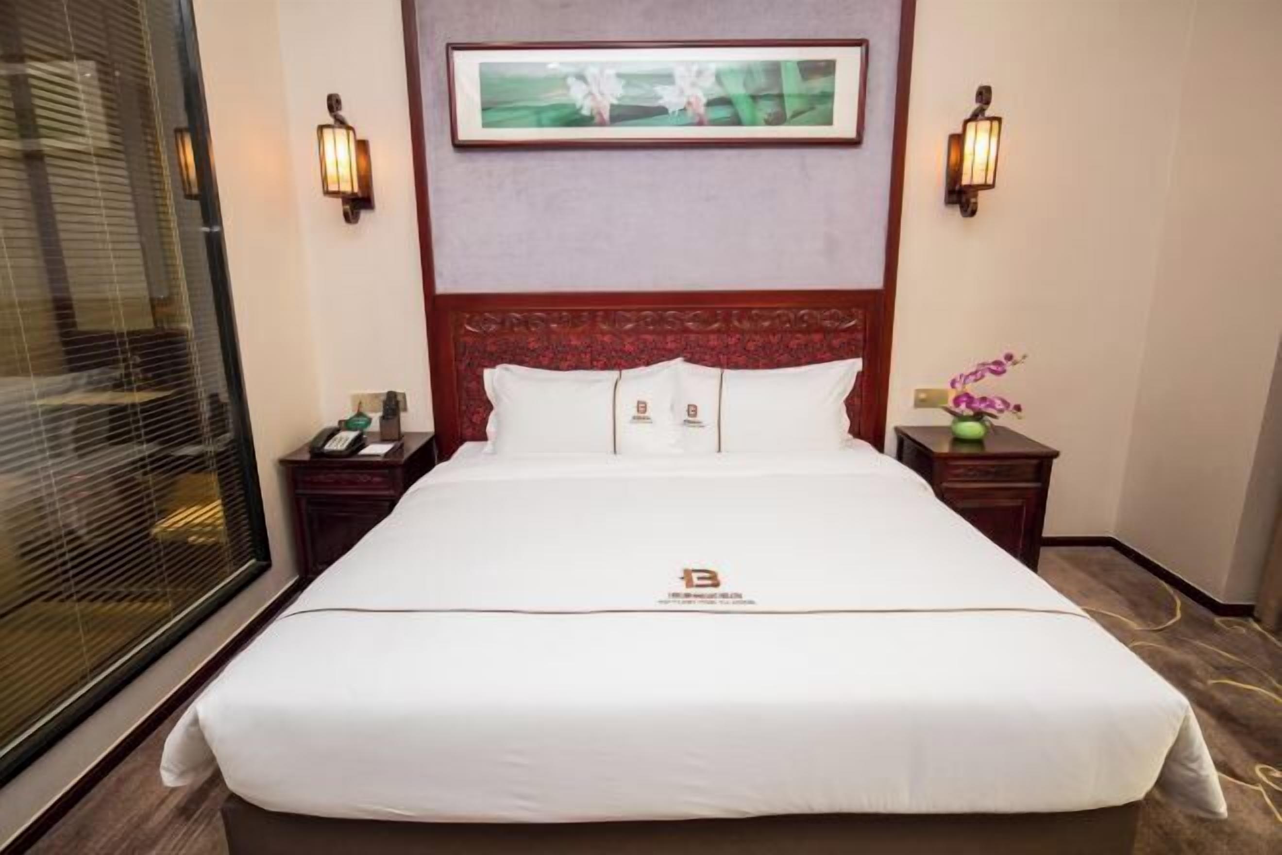 A hotel room with a large bed, two bedside tables, a nightstand, a framed picture above the bed, and a window with blinds.