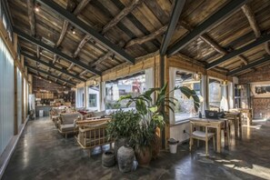 Un interior con techo de madera rústica, una zona de estar central con mesas y sillas, y amplias ventanas que proporcionan luz natural.