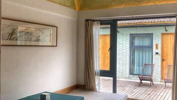 A room with a bed, a table, and a view of an outdoor area with a wooden deck and a brick building.
