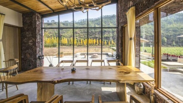 Uma sala de jantar com uma grande mesa de madeira e cadeiras, paredes de pedra e janelas amplas com vista para um vinhedo.