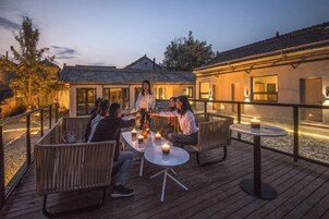 Un grupo de personas sentadas en una terraza de madera con mesas y sillas, con vistas a un edificio con tejado de tejas.