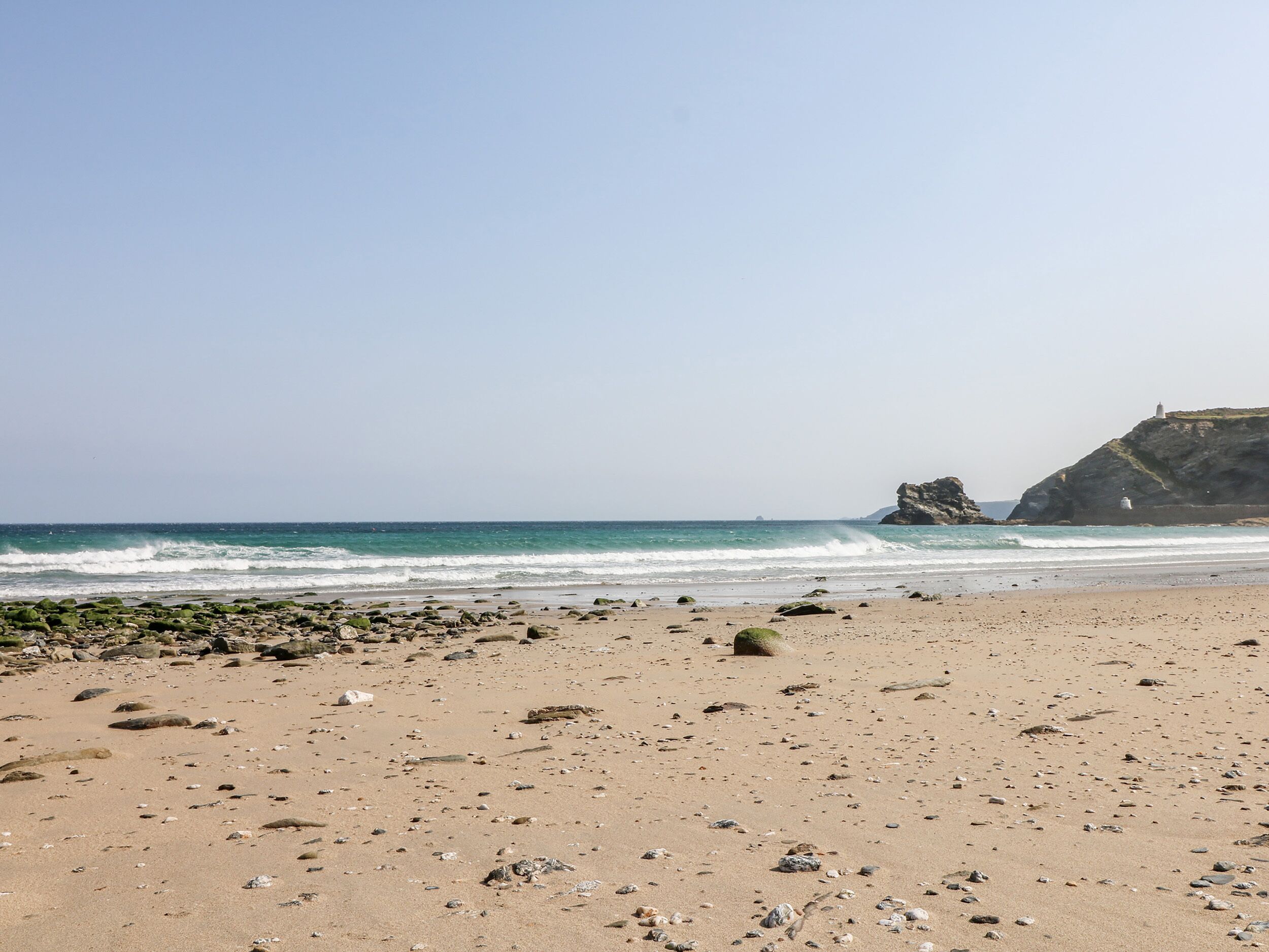 Ein Strand mit felsigem Vordergrund und einem Leuchtturm auf einer Klippe im Hintergrund.