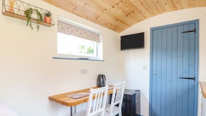 Una cocina compacta con cubierta de madera, sillas blancas, un refrigerador negro, una ventana con persiana estampada y una puerta azul.