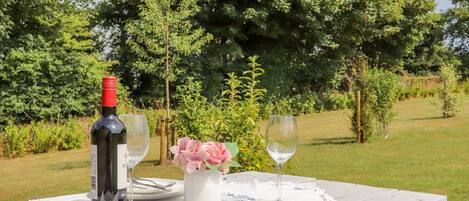 A table set outdoors with a bottle of wine, two glasses, a vase with flowers, and a white tablecloth.