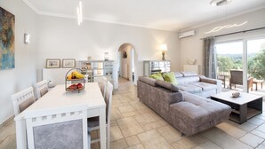 Amplia sala de estar con un sofá seccional gris, una mesa de comedor con sillas, y una zona de cocina con fruteros.