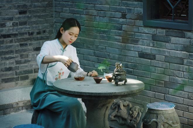 A person pouring tea into a cup on a round table with a decorative stone base, set against a brick wall.