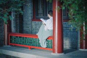 A person sitting on a bench with a red frame, reading a book, in front of a brick building with a window. - Liuyuan Hotel (Tianjin)