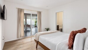Un dormitorio con piso de madera, una cama con ropa blanca y un plaid marrón, un televisor montado en la pared y un ventanal corredero que da a un balcón con zona de comedor.