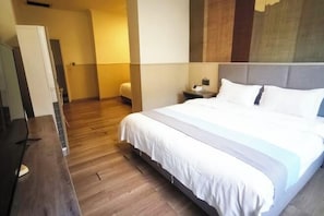 A hotel room with a large bed, wooden flooring, a desk, and a nightstand with a lamp.