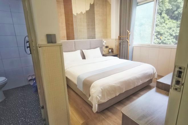 Une chambre d’hôtel moderne avec un grand lit, une table de chevet, une petite lampe de chevet, une fenêtre donnant sur des arbres et une salle de bain visible au fond.