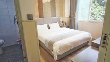 Une chambre d’hôtel moderne avec un grand lit, une table de chevet, une petite lampe de chevet, une fenêtre donnant sur des arbres et une salle de bain visible au fond.