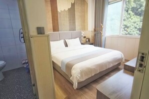 A modern hotel room with a large bed, a bedside table, a small bedside lamp, a window with a view of trees, and a bathroom visible in the background.