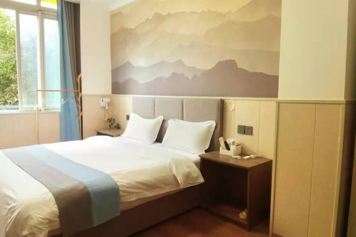 A hotel room with a bed, a nightstand, a wall-mounted lamp, and a window with curtains.