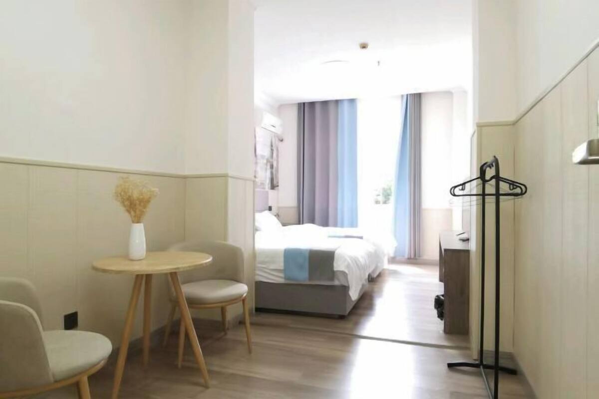 A hotel room with a bed, a small round table with a vase, a chair, and a coat rack.