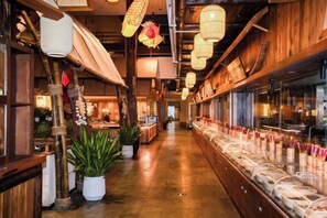 Interior de restaurante com vigas de madeira, lanternas penduradas e um balcão comprido com produtos alimentares.