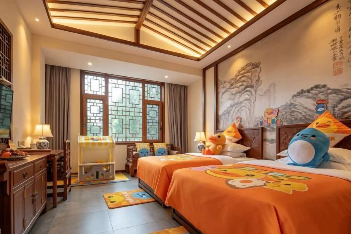 A hotel room with two beds, a desk, and a chair. There are decorative pillows and a wall painting.