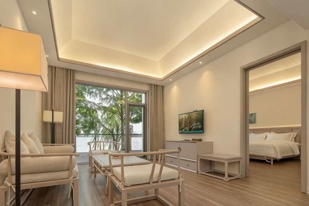 A modern hotel room with a large window, a sofa, a small table, a bed, and a TV.