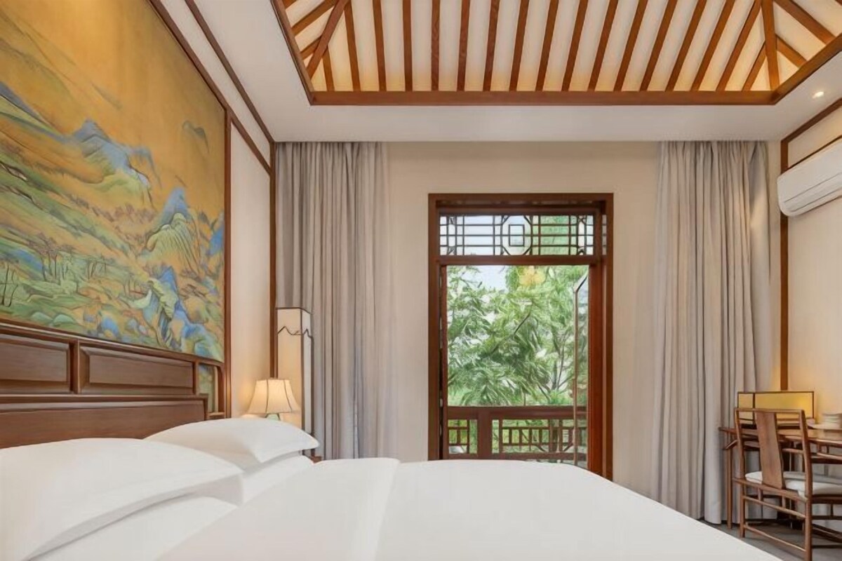 A hotel room with a large bed, a painting on the wall, a window with a view of greenery, and a desk with chairs.