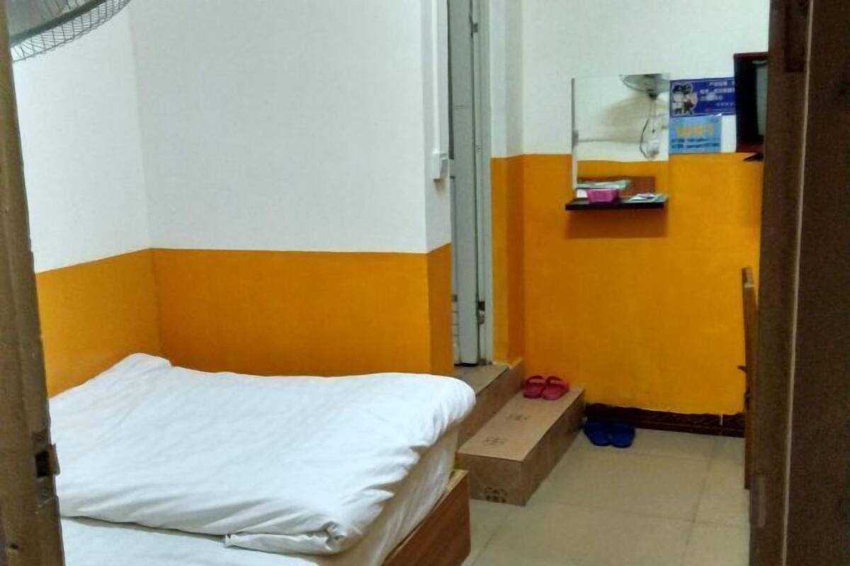 Una habitación pequeña con cama individual, paredes color naranja, un estante con objetos y una puerta que da a otra habitación.