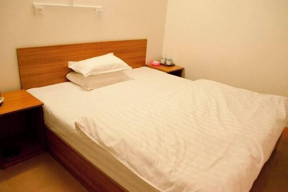 Una cama individual con cabecera de madera, una mesita de noche con lámpara y un enchufe en la pared.