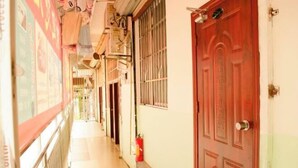 Un couloir étroit avec une porte rouge sur le côté droit, un extincteur accroché au mur et une fenêtre munie de barreaux en métal.